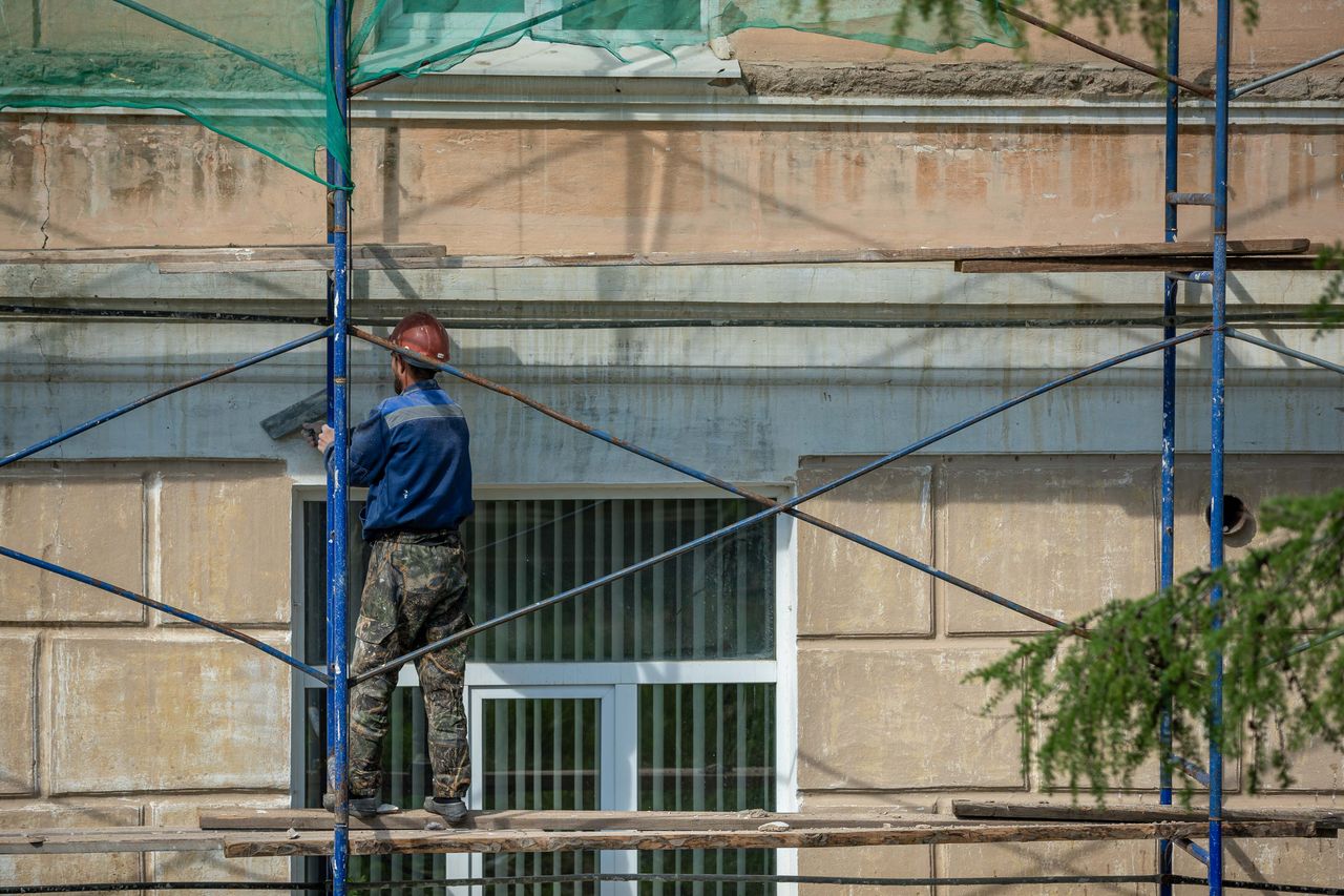 services-02 A construction worker in a hard hat repairs a building facade on scaffolding in an urban setting.